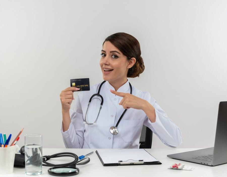 impressed-young-female-doctor-wearing-medical-robe-stethoscope-sitting-desk-with-medical-tools-laptop-holding-pointing-credit-card-isolated_141793-68735
